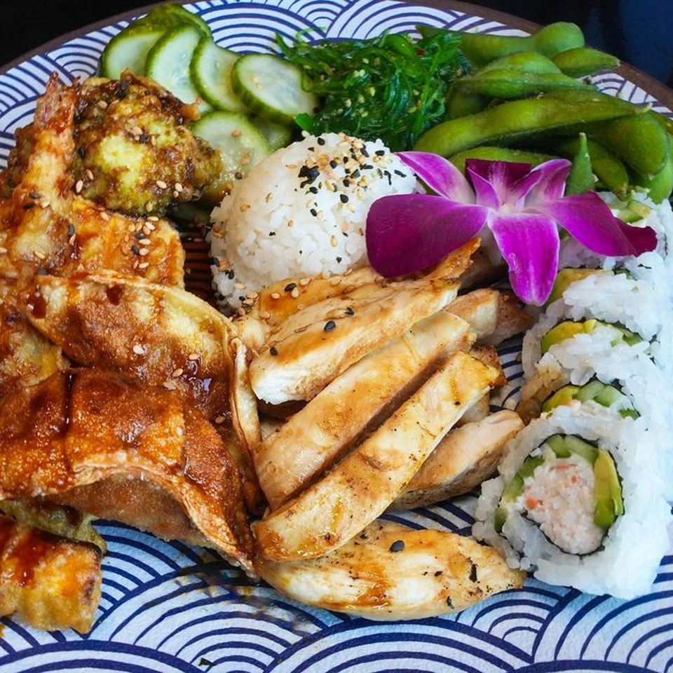 A sushi bento box with California rolls, chicken, dumplings, rice, edamame, seaweed salad, and cucumbers.