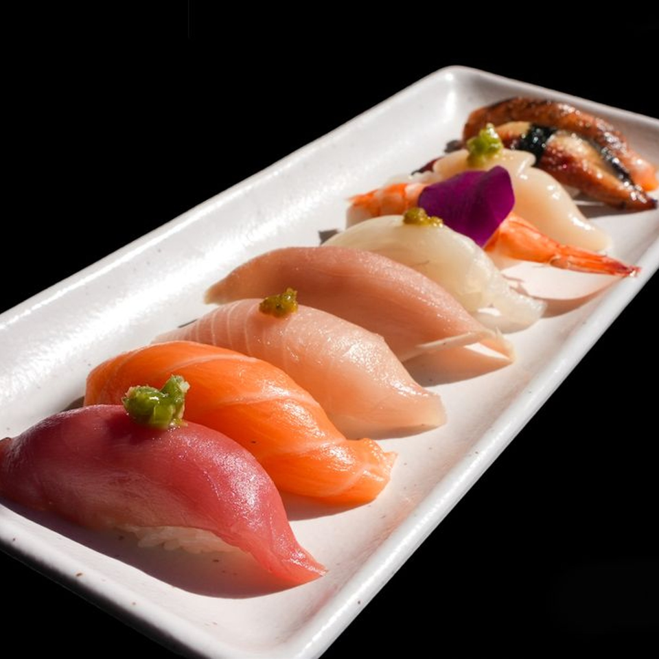 A white rectangular plate holds a variety of colorful sushi nigiri against a dark background.