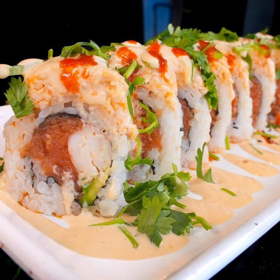 Sushi rolls topped with creamy sauce, red drizzle, and fresh cilantro on a white plate.